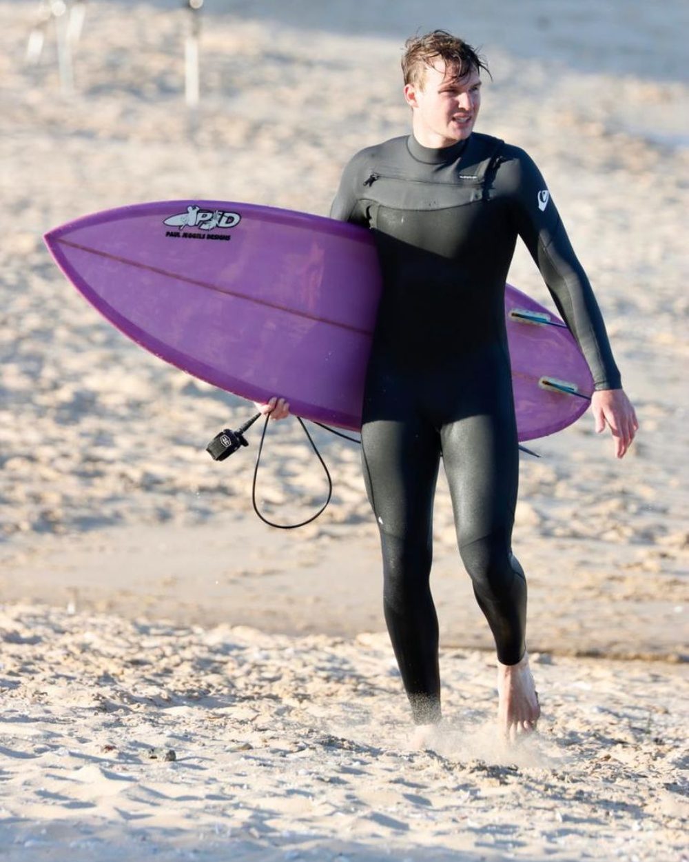 A surfer in a wetsuit walking along the beach carrying his purple custom surfboard from Paul Jeggels Designs.