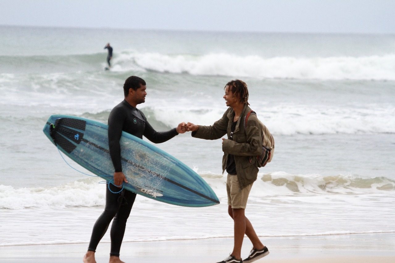 Two surfers meeting on the beach and bumping fists, one of them in a wetsuit with a well used blue board under his arm.