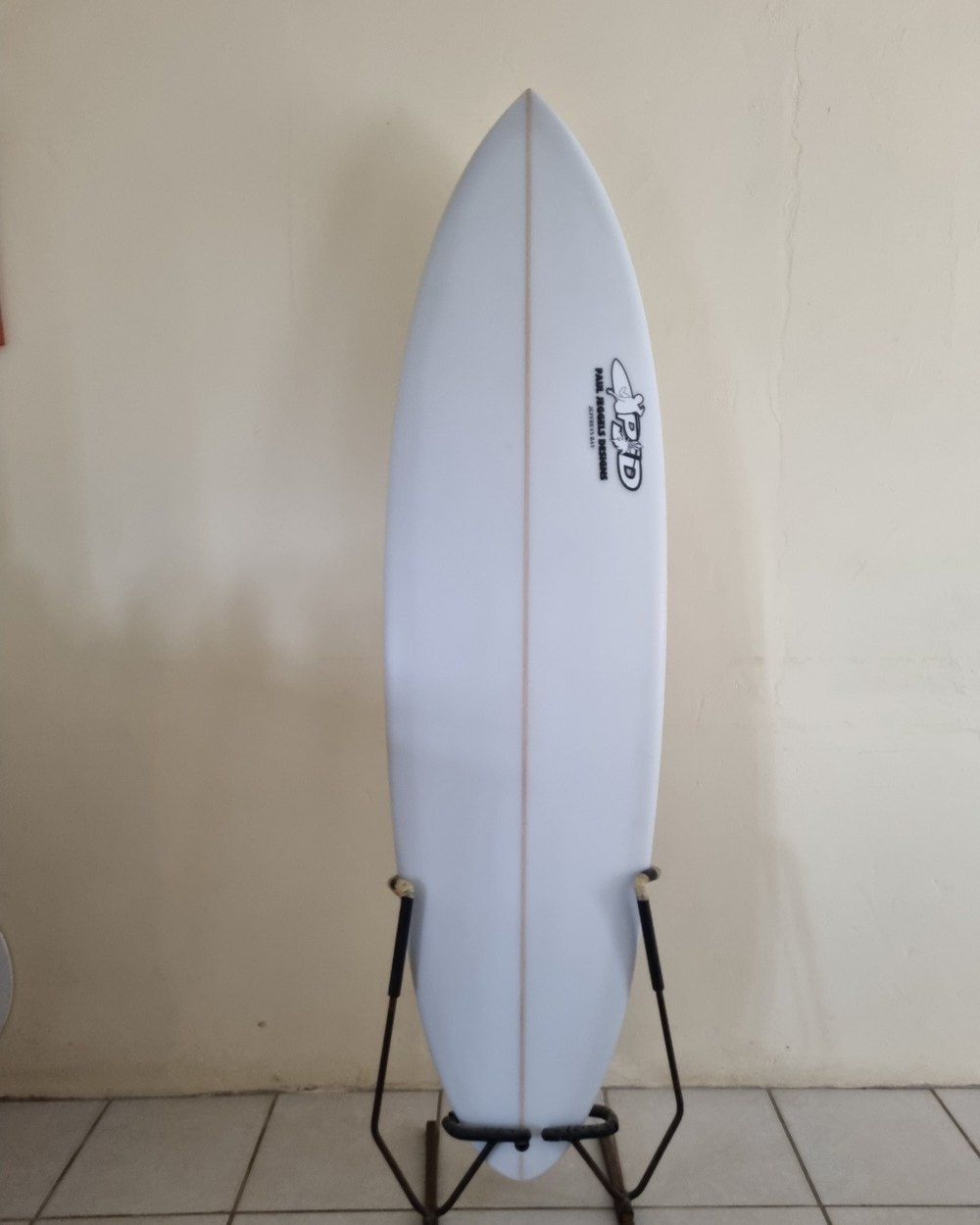 A finished Paul Jeggels Designs surfboard on a stand against a wall, the maker's PJD logo and Jeffreys Bay lettering on the deck and the stringer line running down the centre.