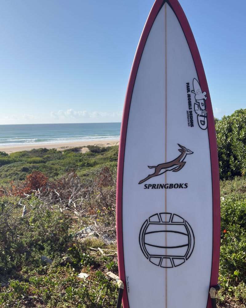 A finished Paul Jeggels Designs surfboard on a stand above the beach at Jeffreys Bay, the PJD logo and Springbok crest on the deck
